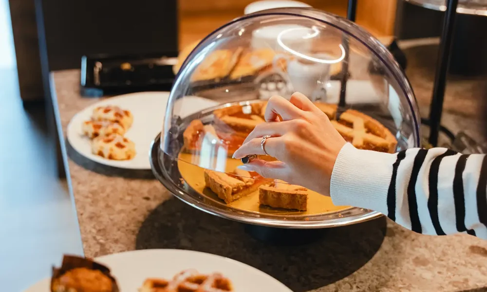 Una mano prende una fetta di dolce da un vassoio coperto su un tavolo da buffet, circondata da vari dolci e frutta.