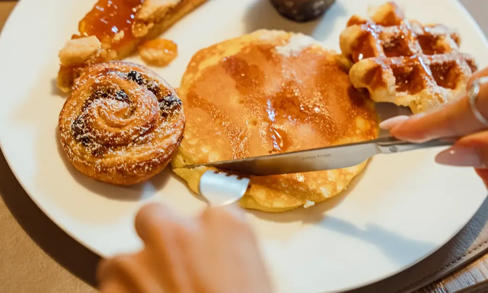 Persona che taglia una frittella su un piatto con vari dolci per colazione.