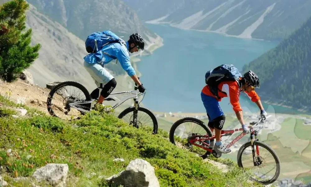 Due ciclisti in mountain bike percorrono un sentiero montano con vista su un lago alpino.