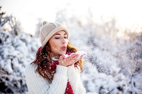 Donna con berretto che soffia neve tra le mani in un paesaggio invernale.